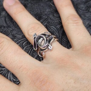 May include: A silver-colored ring featuring a detailed snake design. The snake is coiled around the finger, with intricate scales and a textured surface. The ring is a statement piece, perfect for those who appreciate unique jewelry.