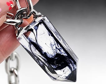 以下が含まれることがあります： 内部に渦巻く濃い紫と黒のインクのような模様が入った、透明な六角形の樹脂製ペンダント。ペンダントは太い光沢のあるシルバー調の金属チェーンに取り付けられており、大胆で幾何学的なアクセサリーとなっています。