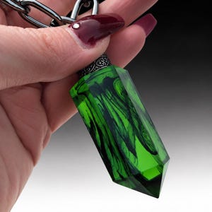 May include: A pendant shaped like a faceted crystal point, featuring a translucent green resin body with swirling black ink-like patterns. The top is capped with a decorative silver-toned metal bail attached to a thick, dark gunmetal chain link.