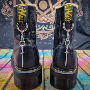 Könnte beinhalten: Schwarze Lederstiefel mit gelben Nähten und einem silbernen Schwertanhänger, der an den Schnürsenkeln befestigt ist. Die Stiefel haben eine dicke Plateau-Sohle und einen Reißverschluss an der Seite.