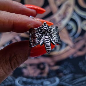 May include: A silver ring featuring an intricate design of a moth with a skull on its head. The ring is worn on a finger with orange nail polish.