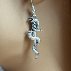 May include: A silver-toned dangle earring featuring a detailed metal snake coiled around a straight sword. The snake has visible scales and an open mouth, while the sword has a simple crossguard. The earring hangs from a standard silver hook.