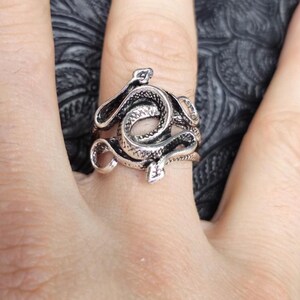 May include: A silver-toned ring featuring a coiled snake design. The ring has intricate detailing, with the snake's scales and head clearly visible. The ring is worn on a finger, showcasing the jewelry's design and craftsmanship.