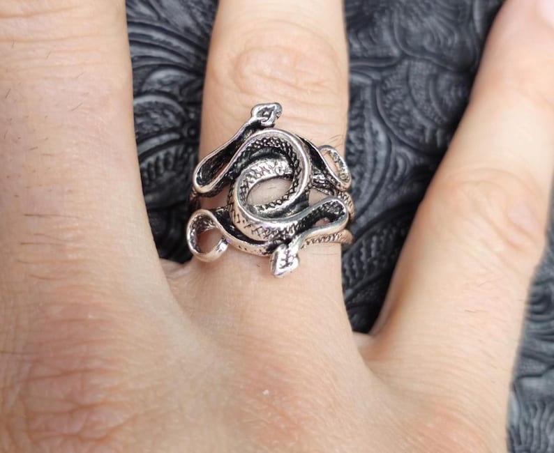May include: A silver-toned ring featuring a coiled snake design. The ring has intricate detailing, with the snake's scales and head clearly visible. The ring is worn on a finger, showcasing the jewelry's design and craftsmanship.
