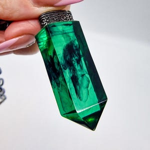 May include: A translucent emerald green pendant shaped like a faceted crystal point. The interior features dark, swirling smoke-like patterns suspended within the resin. It is attached to a decorative, dark metallic cap with an intricate filigree design.