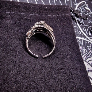 May include: Silver-toned ring with an open design, featuring a snake motif. The ring has intricate detailing and a small, clear stone. The ring is set against a dark, textured background, with dried rose buds and a decorative cloth visible.