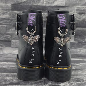 Könnte beinhalten: Ein Paar schwarze Lederstiefel mit silberfarbenen Totenkopf-Motten-Anhängern an den Fersenschlaufen. Die Stiefel haben eine violette AirWair-Stickerei auf den Schlaufen, silberne Schnallen, gelbe Nähte und den aufgedruckten Text ARE IN.