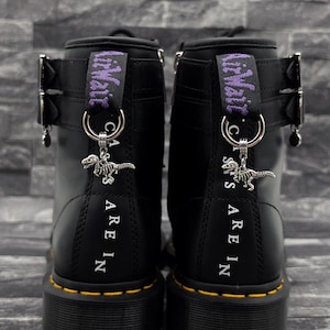 Könnte beinhalten: Ein Paar schwarze Lederstiefel mit gelben Nähten an den Sohlen, silbernen Metallschnallen und violetten Fersenschlaufen mit der Aufschrift AirWair. Jeder Stiefel hat einen silbernen Dinosaurierskelett-Anhänger an der Schlaufe.