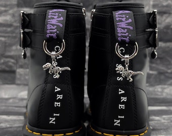 Puede incluir: Un par de botas de cuero negro con costuras amarillas en las suelas, hebillas de metal plateado y tiradores de talón morados con la etiqueta AirWair. Cada bota tiene un dije de esqueleto de dinosaurio plateado en la trabilla.