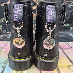 May include: Pair of black leather boots with yellow stitching and silver hardware. Each boot has a purple and black fabric loop with the text "McWatt". Silver charms with a moth design hang from the loops. The text "MADE IN" is printed on the back of each boot.