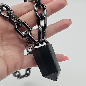 Könnte beinhalten: Eine Halskette mit einer dicken, glänzend schwarzen Kette aus großen ovalen Gliedern. Ein glatter, facettierter schwarzer Steinanhänger in Form eines spitzen hexagonalen Prismas hängt in der Mitte. Das gesamte Stück hat ein einheitliches Finish.