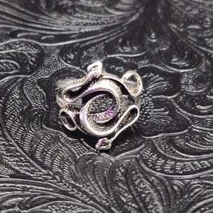 May include: A silver-colored ring featuring a detailed snake design. The snake is coiled and intricately crafted, with visible scales and curves. The ring is set against a textured, dark background, highlighting the metallic sheen of the jewelry.