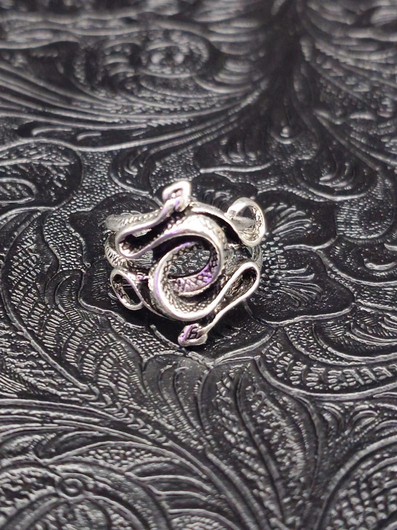 May include: A silver-colored ring featuring a detailed snake design. The snake is coiled and intricately crafted, with visible scales and curves. The ring is set against a textured, dark background, highlighting the metallic sheen of the jewelry.