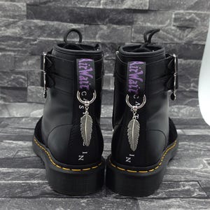 Könnte beinhalten: Ein Paar schwarze Lederstiefel mit doppelten Schnallenriemen, gelben Rahmennähten und violetten AirWair-Fersenschlaufen. Jeder Stiefel ist mit einem silberfarbenen Federanhänger versehen, der an einem Metallring an der Fersenlasche befestigt ist.