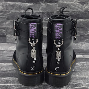 Könnte beinhalten: Ein Paar schwarze Stiefel mit silbernen Schnallen, gelben Nähten an der Sohle und violetten AirWair-Fersenschlaufen. Jeder Stiefel hat einen silbernen Metallanhänger an einem Ring sowie den vertikal aufgedruckten Text AirWair und A S T R E I N.