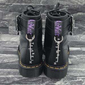 Könnte beinhalten: Ein Paar schwarze Lederstiefeletten mit silbernen Schwertanhängern an den Fersenschlaufen. Die Stiefel haben violetten AirWair-Text auf den Schlaufen, silberne Schnallen an den Seiten, gelbe Nähte entlang der Sohlen und weißen Text auf der Rückseite.