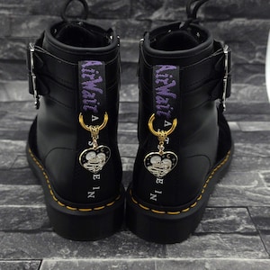 May include: A pair of black leather boots featuring purple AirWair heel loops and silver-tone buckles. Each boot is adorned with a gold-tone ring holding a heart-shaped charm that depicts two skeletons embracing, with white and black enamel details.