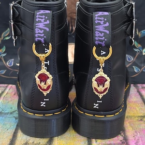 May include: A pair of black leather boots featuring yellow stitching, silver buckles, and purple AirWair heel loops. Each boot is adorned with a gold-toned pendant featuring a red oval frame and a central bat motif, suspended from a gold ring.