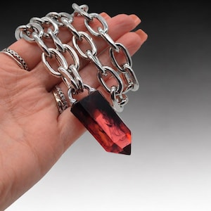 May include: A necklace featuring a thick, polished silver-tone metal chain with large oval links. A faceted, translucent deep red crystal pendant with dark, smoky inclusions hangs from the center of the chain, secured by a silver-tone metal bail.