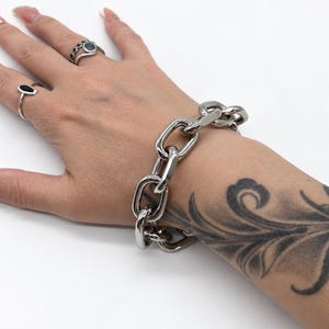 May include: A chunky silver-toned metal chain bracelet featuring large, polished oval links. The bracelet is worn around the wrist, showcasing a high-shine, reflective surface and a substantial, heavy-duty link design that connects seamlessly.
