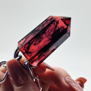May include: A faceted, hexagonal crystal-shaped pendant made of translucent red resin with swirling black ink-like patterns inside. The pendant is attached to a polished silver-toned metal chain link, creating a bold and geometric accessory piece.