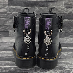 Könnte beinhalten: Ein Paar schwarze Lederstiefel mit zwei silbernen Schnallen an der Seite, gelben Nähten an der Sohle und einer violetten Fersenschlaufe mit dem Text AirWair. Ein silberner Sonnen- und Mondanhänger hängt an einem Ring an jeder Schlaufe.