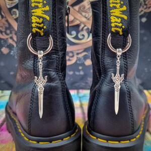 May include: A pair of black leather boots featuring yellow stitching along the soles and black heel loops with yellow AirWair text. Each boot is adorned with a silver-toned metal sword charm attached to a circular ring on the heel loop.
