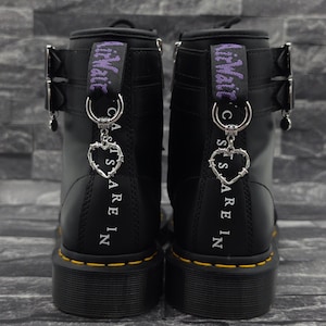 May include: A pair of black leather boots featuring yellow welt stitching, silver-toned buckles, and purple heel loops. Each boot is adorned with a silver-coloured heart-shaped charm made of barbed wire, hanging from a metal ring on the heel tab.