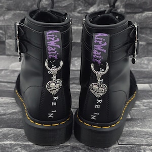 Könnte beinhalten: Ein Paar schwarze Stiefeletten mit gelben Nähten an der Sohle, silbernen Schnallen und violetten AirWair-Fersenschlaufen. Jeder Stiefel hat einen silbernen herzförmigen Anhänger mit zwei Skeletten und dem vertikal aufgedruckten Text RE IN.