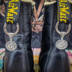 May include: A pair of black leather boots with a yellow and black striped strap and a silver charm attached to each strap. The charms feature a silver design of a wolf howling at the moon.