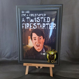 Könnte beinhalten: Ein gerahmter Druck eines Mannes mit einem schockierten Ausdruck, mit dem Text "Oh God I'm a Firestarter A Twisted Firestarter" darüber. Der Druck steht auf einem kleinen Holzstaffelei und hat einen schwarzen Rahmen mit einem schwarzen Katzenaufkleber unten rechts.