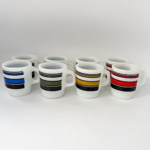 May include: A set of eight white milk glass mugs featuring horizontal stripes in blue, olive green, yellow, and red. Each mug has a side handle and a stackable design, with black accent lines bordering the coloured bands around the exterior.