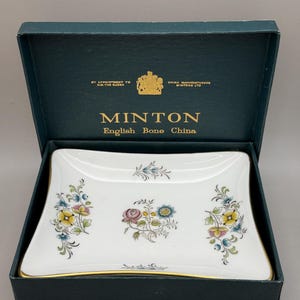 May include: A rectangular white Minton English Bone China dish with gold trim, featuring colourful floral patterns in the centre and on the sides. The dish is presented in a dark teal box with gold text reading By Appointment to H.M. The Queen.