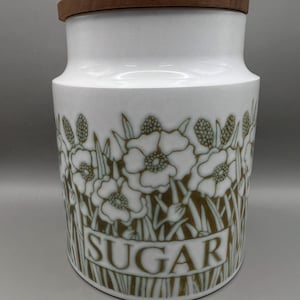 May include: White ceramic sugar jar with a wooden lid. The jar has an olive green floral pattern and the word "SUGAR". The jar is suitable for storing sugar.