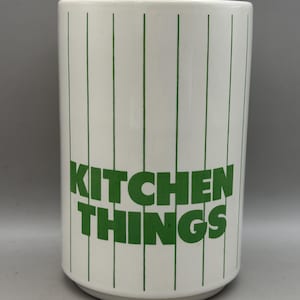 May include: A white cylindrical ceramic utensil holder featuring thin vertical green pinstripes and the text KITCHEN THINGS printed in bold green capital letters across the centre of the exterior surface of the container.