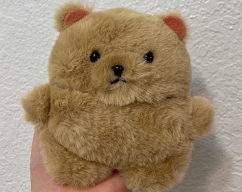 May include: A small, brown plush bear with a round body and a flat face. The bear has black eyes, a black nose, and a small, black mouth. The bear's ears are a light pink color.