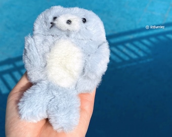 May include: A small, plush seal toy with light blue fur and a white belly patch. The seal has two black bead eyes and a small black nose. The text litfurries is printed in white on the right side of the plush toy, which is held in a palm.