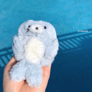 May include: A plush, light blue seal toy with a white belly and black eyes and whiskers. The toy is held in a hand against a blue background. The text "litfurries" is in the upper right corner.