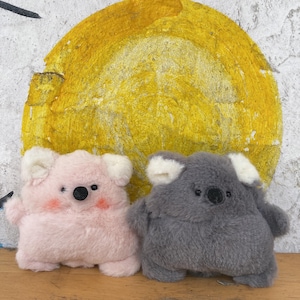May include: Two plush koala toys. One is pink with rosy cheeks, the other is grey. Both have white ears and black eyes and noses. They are set against a background of a yellow circle on a textured wall.