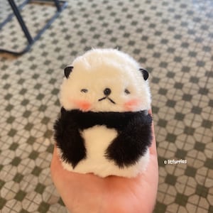 May include: A small white plush panda bear wearing a black scarf. The panda has black eyes and a black nose. The panda is sitting on a hand.