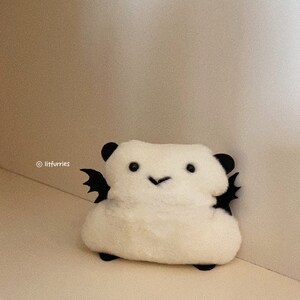 May include: A white plush toy with black wings, ears, and feet. The soft toy has a smiling face with black eyes and a small, black stitched mouth. The product is a handmade item.