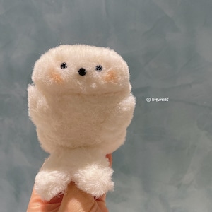 May include: A fluffy white plush toy shaped like a small creature with a rounded body, small black eyes, and rosy cheeks. The toy has small arms and legs, and is held against a light blue background. The text "@litfurries" is in the bottom right corner.