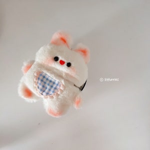 May include: A white plush toy shaped like a bunny with pink accents and blue and white checked fabric on its chest. The toy has black eyes and a red nose.