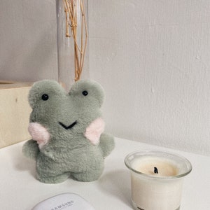 以下が含まれることがあります： ピンクのアクセントが付いた緑色のぬいぐるみのかえるの玩具。かえるは笑顔で黒い目です。玩具は白い表面に、透明なガラスのキャンドルホルダーと白いキャンドルの隣に置かれています。