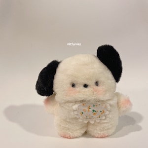 Könnte beinhalten: Weißes Plüsch-Hunde-Spielzeug mit schwarzen Ohren und einer Blumenschürze. Das Spielzeug hat ein kleines, rundes Gesicht mit schwarzen Augen und rosa Wangen.