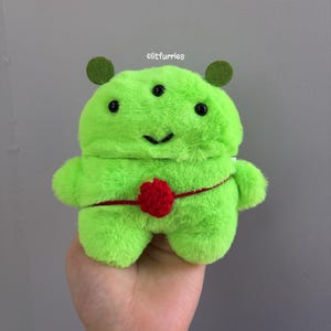 May include: A plush green alien toy with three eyes, two small green antennae, and a red crocheted detail around its midsection. The toy has a smiling face and is made of soft, fuzzy material. The text "@litfurries" is in the top right corner.