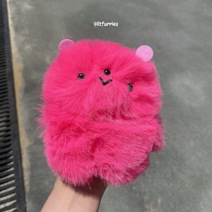May include: A vibrant pink, fluffy creature with small, round ears and black embroidered eyes and a smile. The plush toy has a soft, furry texture and is designed to resemble a cartoon character. The text "olitfurries" is visible in the top right corner.