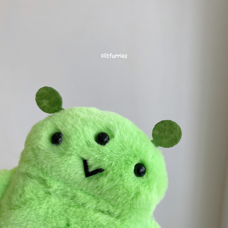 May include: A bright lime green plush toy with a soft, fuzzy texture. The creature features two small, rounded dark green ears, three black bead eyes, and a simple black stitched V-shaped mouth. The text litfurries is printed above the toy.