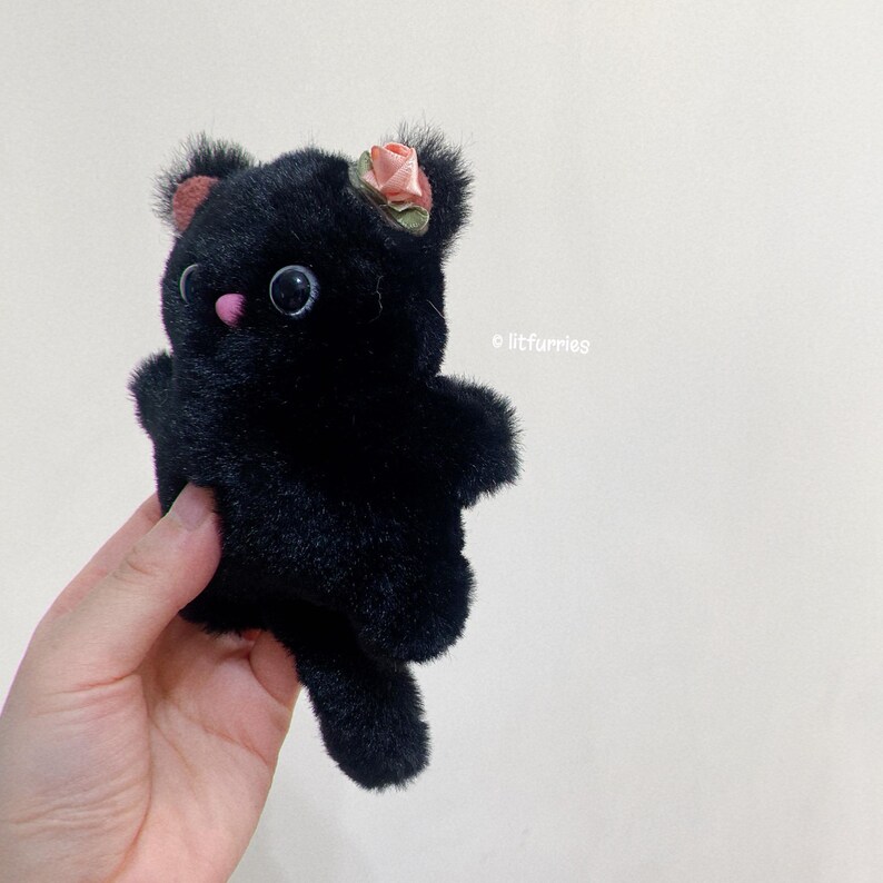 May include: A black plush toy cat with a pink flower on its head. The cat has large black eyes and a pink nose. The toy is being held in a hand.