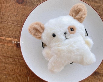 May include: A white plush toy dog with brown ears and a brown spot on its face. The toy is lying on a white plate with brown speckles.